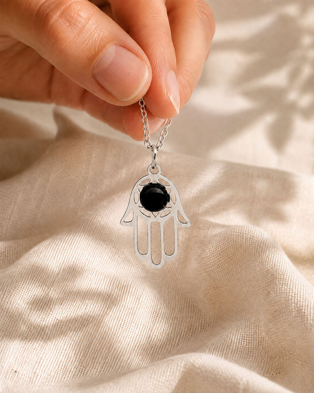 Mano sosteniendo el colgante Mano de Luz (Hamsa) con obsidiana, cadena cayendo entre los dedos sobre lino crema