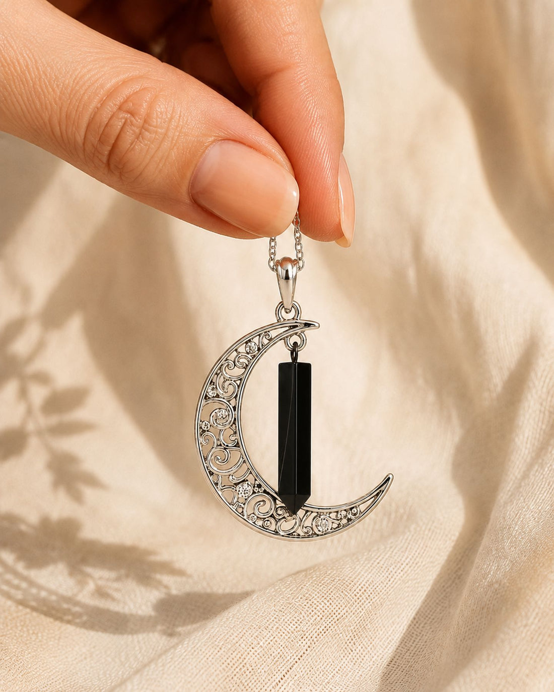 Mano sosteniendo el colgante Luna Protectora con punta pulida de turmalina negra, cadena cayendo entre los dedos sobre lino crema